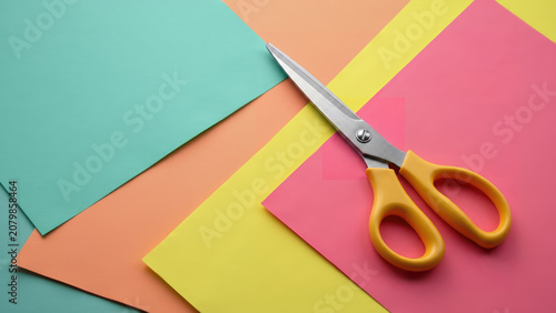 Yellow scissors rest diagonally on overlapping sheets of pastel teal, peach, yellow, and pink paper in a vibrant, minimalist flat lay.