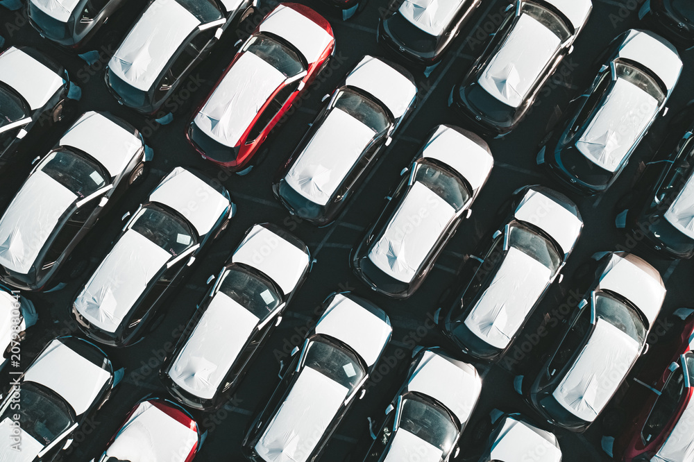 Fototapeta premium Aerial top down view of new cars parked in car parking lot. Car dealer parking lot full of new automobiles. New cars lined up for import and export business.