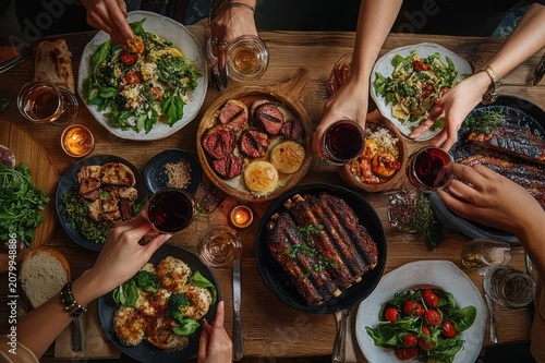 Warm convivial dinner with hands reaching over a wooden table laden with grilled ribs, steak medallions, salads, roasted vegetables, crusty bread, wine and candlelight