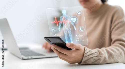 Woman holding smartphone with virtual human body and vital signs interface on screen in office with laptop