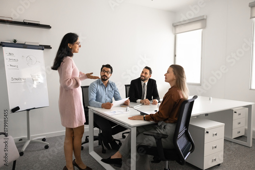 Businesswoman stands near flipchart presenting strategy to colleagues seated at conference table, explaining marketing plan. Business presentation, discussion, collaborative decision making in office