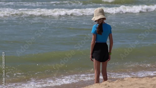 Pretty little girl in a swimsuit, sun glasses and hat relaxing on the beach near sea. Smiling cute little girl on vacation. Adorable girl in a big straw hat looks at the sea. Summer travel concept