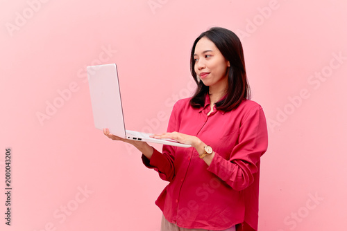 Successful young Asian businesswoman smiling while working on her laptop, standing against a pink isolated background.