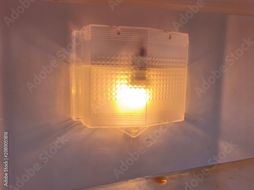 Interior view of an empty refrigerator compartment with glowing yellow light bulb, close up of a bright warm fridge lamp behind textured plastic cover casting shadow on white freezer wall