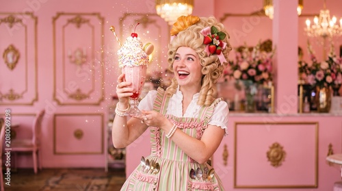 Cheerful woman in pink striped rococo costume with pastel wig decorated with croissant and berries, holding oversized whimsical milkshake dessert at colorful candy bakery setting.