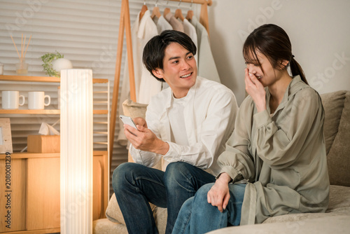 Bild auf Leinwand A young Asian couple chatting while looking at their smartphones on the sofa in their living room