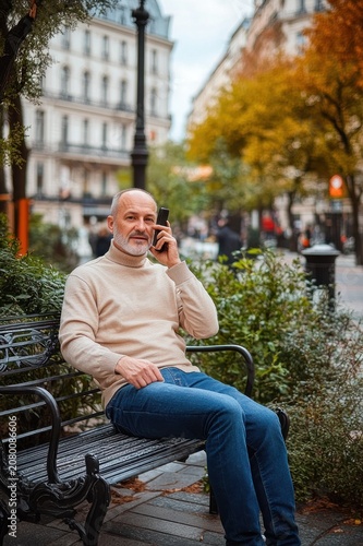 Man sitting on park bench in city street during autumn talking on smartphone, relaxed and contemplative urban scene
