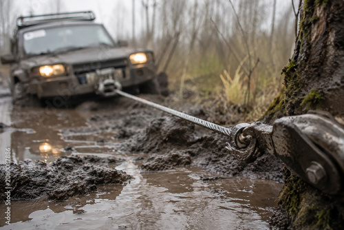 Off-road vehicle stuck in mud being pulled by a winch cable attached to a tree on offroad background.