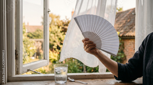 Woman hand holding paper fan by open window with glass of water and spoon for summer cooling background and lifestyle banner