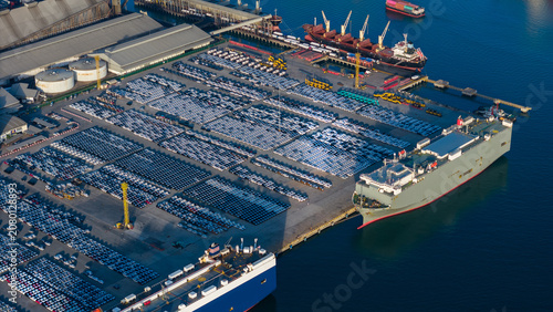 Картината върху платно roll-on roll-off (RoRo) vessel docks at a major industrial port facility, surrounded by thousands of new vehicles staged for global export alongside busy container shipping terminals
