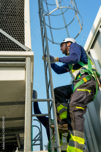 Engineer ascending cooling tower ladder. Rooftop HVAC system maintenance, industrial worker inspecting cooling unit for optimal performance.