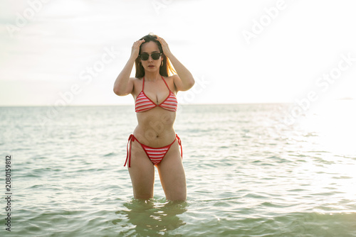 Fotografía Beautiful young woman in a red striped bikini and sunglasses standing in the sea water holding her hair
