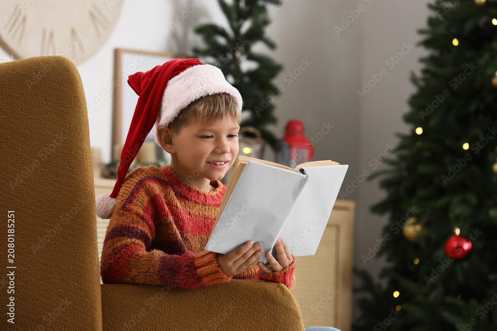 Obraz premium Cute little boy with Santa hat reading Christmas story in armchair at home