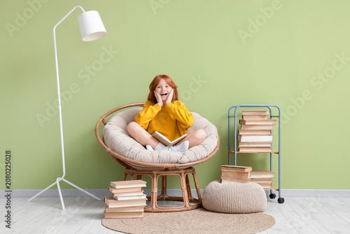 Cute redhead girl with book in armchair near green wall