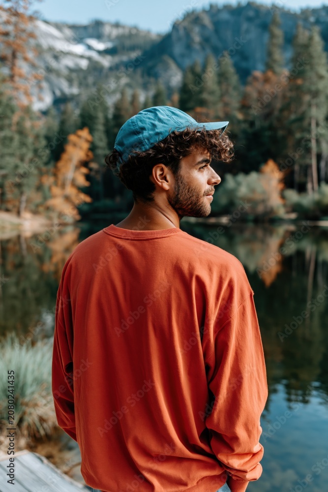 Fototapeta premium Man in an orange sweatshirt and teal cap standing on a pier by a forest lake with mountains in the background for outdoor lifestyle and travel use