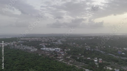 Aerial view of houses in Old Goa surrounded by lush greenery during the pre-monsoon season, with the Mandovi River in the background and a dramatic sunset beneath cloudy skies.
