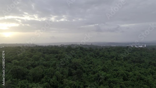 Aerial view of houses in Old Goa surrounded by lush greenery during the pre-monsoon season, with the Mandovi River in the background and a dramatic sunset beneath cloudy skies.