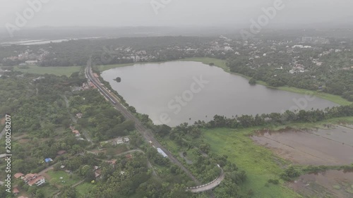 Aerial view of a train at Karmali Railway Station near Carambolim Lake in Old Goa, surrounded by lush greenery, coconut palms, and trees beneath cloudy monsoon evening skies.