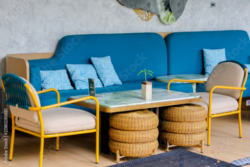 Blue sofa, yellow chairs, woven stools, and a marble table. Cozy cafe corner, bright and inviting. Light gray walls, minimalist decor. Perfect for casual meetings or relaxed gatherings