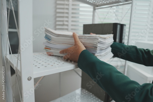 Photos An office employee standing behind a light wooden desk uses both hands to lift and vertically align a heavy stack of white document