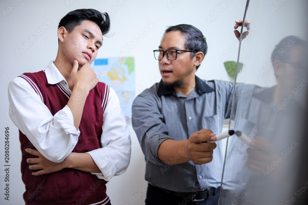 Obraz premium male Teacher with Glasses Explaining Lesson on Whiteboard to Thoughtful Young male Student in Class