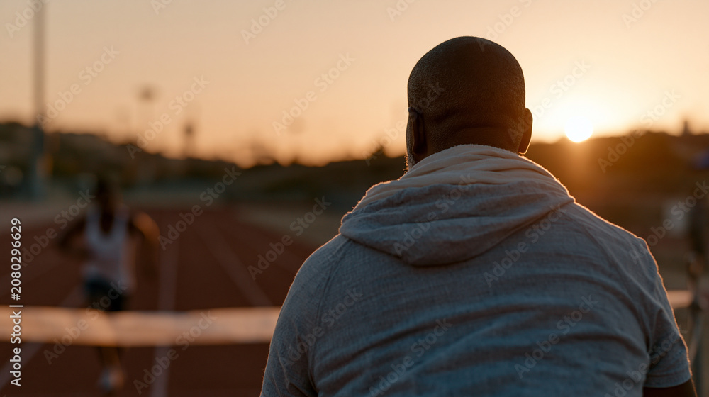 Obraz premium Ghanaian male track coach watching a sole runner burst through a paper finish line tape on an empty outdoor track at sunrise, low angle perspective emphasizing speed and forward