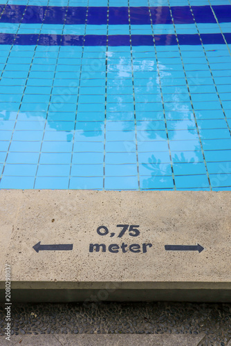 Clear blue pool tiles reflect sky above. Concrete edge shows 0.75 meter marker with arrows. Pool depth guide helps swimmers measure distance. Ideal for swimming facility or training center