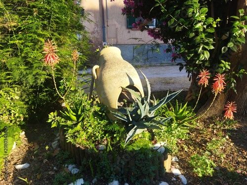 Tinaja de terracota decorativa rodeada de plantas de aloe vera en flor y vegetación en un jardín al atardecer, Toscana, Italia.