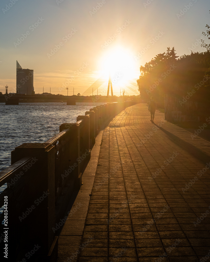 Obraz premium Golden sunset over a riverside promenade with a cable-stayed bridge, warm sun rays and a lone walking silhouette creating a peaceful urban evening atmosphere.