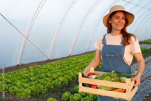 Woman farmer in straw hat holding crate of fresh vegetables inside a greenhouse