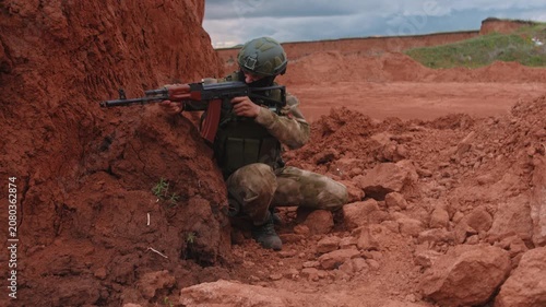 Camouflaged soldier kneels in red clay ravine aiming rifle with focused posture using earthen cover during military training outdoors under cloudy sky side view
