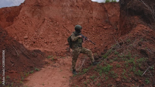 Soldier in tactical gear aiming rifle while advancing across steep red clay slope, alert stance on rugged desert terrain with embankment and overcast light, with soldier, rifle, aiming, tactical
