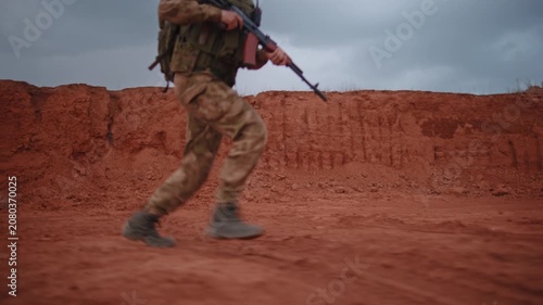 Blurred adult man in camouflage uniform and cap walks across red desert ground with rifle at chest level beside rugged clay embankment under gray overcast sky, low wide outdoor view