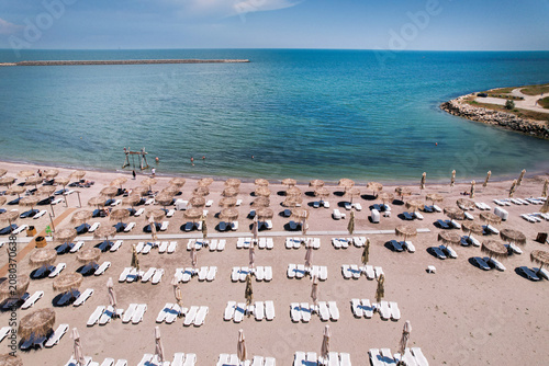 Aerial top down view of an organized sandy beach resort with rows of straw umbrellas and white sun loungers near the sea. Aerial top down view of rows of straw umbrellas and sun loungers on a sandy be