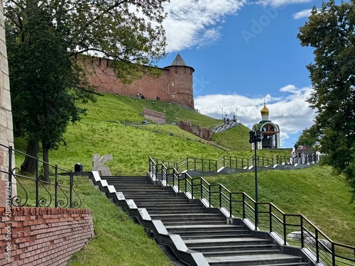 Photos visitors on steps of nizhny novgorod kremlin in peaceful and historical outdoor daytime scene with brick wall, steps, railing, bell tower, and trees under clear sky and greenery