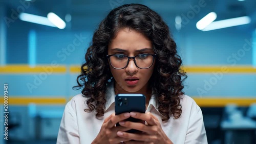 Young Woman with Glasses Looking at Smartphone with a Concerned Expression in a Modern Office Environment.