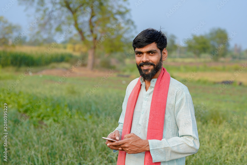Fototapeta premium young indian farmer using smartphone standing at green agricultural field