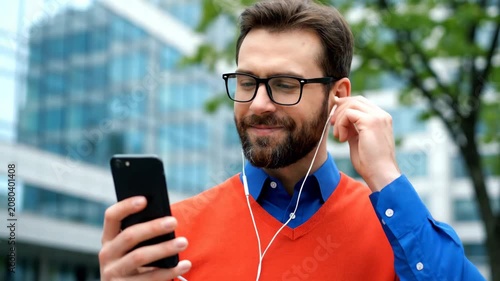 Smiling man in glasses and orange vest using smartphone with earphones outdoors in a city setting.