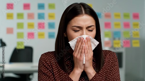 Young woman suffering from a cold, sneezing into a tissue in an office environment, experiencing allergy symptoms.