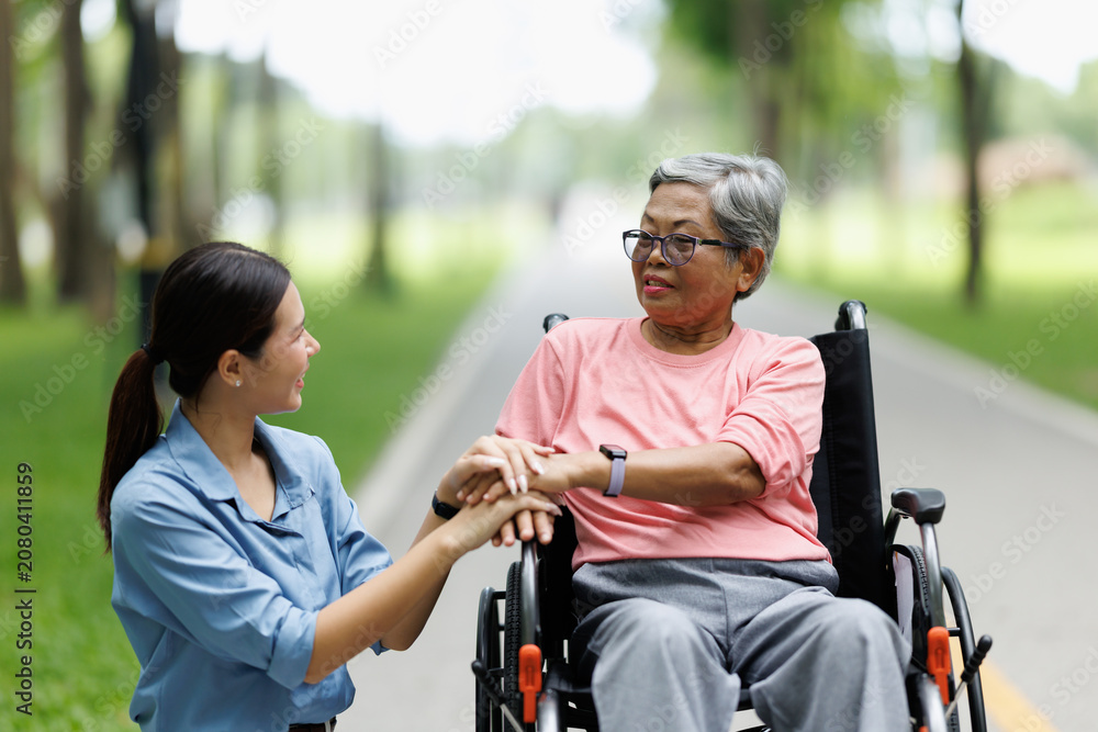 Obraz premium Young caregiver offering compassionate support to a senior woman in a wheelchair, gently holding her hands, symbolizing care, empathy, and assistance during an outdoor park activity