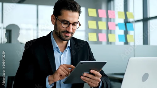 Young businessman with glasses using a digital tablet in a modern office, working on a project with sticky notes on a glass wall in the background.