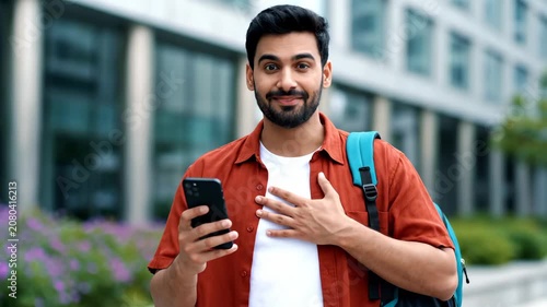 Happy young Indian man holding smartphone and looking at camera with hand on chest, standing outdoors in city.
