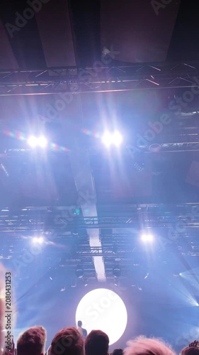 Vertical footage of bright stage lights flaring over a concert crowd, a glowing round screen shines behind the audience as beams cut through haze inside the dark venue