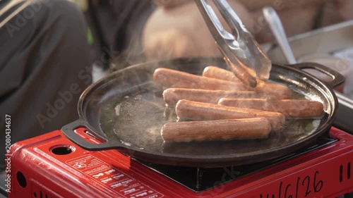 Sausages sizzle on a hot cast-iron griddle over a portable gas stove, with steam rising as they are turned using metal tongs during outdoor cooking.