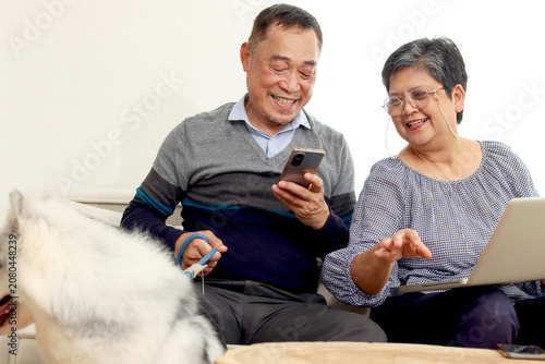 Happy Asian elderly couple sit on sofa in living room with Siberian Huggy dog pet, mature woman uses laptop computer, retired man uses mobile phone. Senior lover relaxes at home, retirement lifestyle.