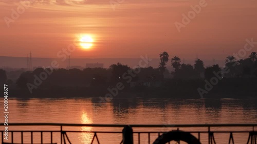 A beautiful golden sunrise over the Nile River in Edfu, Egypt, viewed from a river cruise boat with palm trees and silhouettes on the shore.