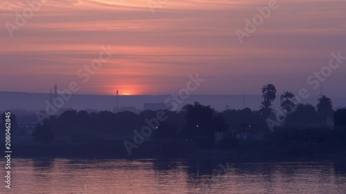 A beautiful sunrise over the Nile River in Edfu, Egypt, featuring silhouettes of palm trees, buildings, and a mosque minaret against a colorful sky.
