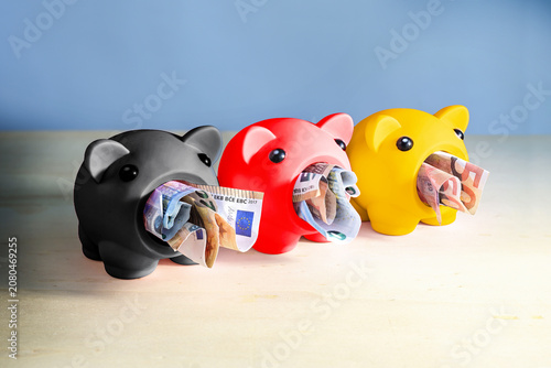 Tableau sur toile Three piggy banks in the German colors, black, red, and gold, devouring euro ban