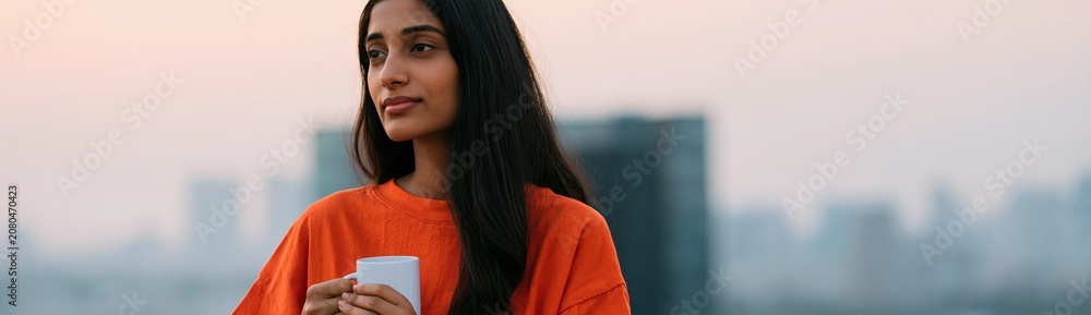 Naklejka premium Young woman holding a mug on a rooftop at sunrise with soft city skyline for lifestyle background banner and calm morning design