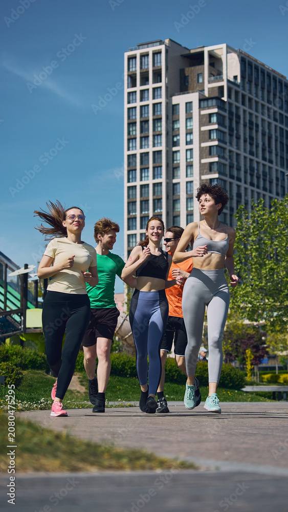 Obraz premium Group of young friends jogging together through modern city park during outdoor fitness workout. Concept of healthy lifestyle, active recreation, cardio exercise, teamwork and urban wellness.